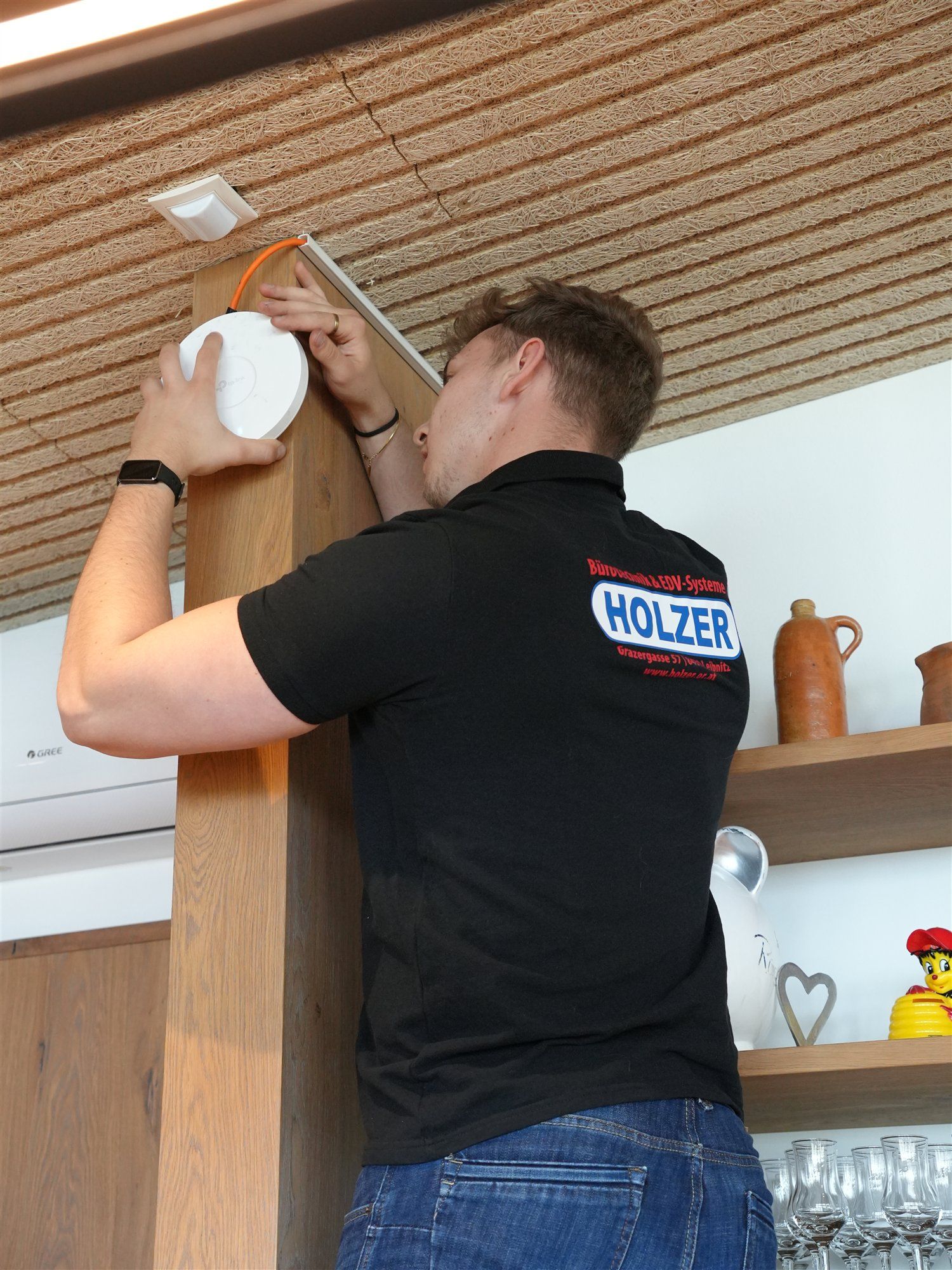 Holzer-Techniker hält im Profil einen WLAN-Access-Point an eine Holzsäule unter der Decke