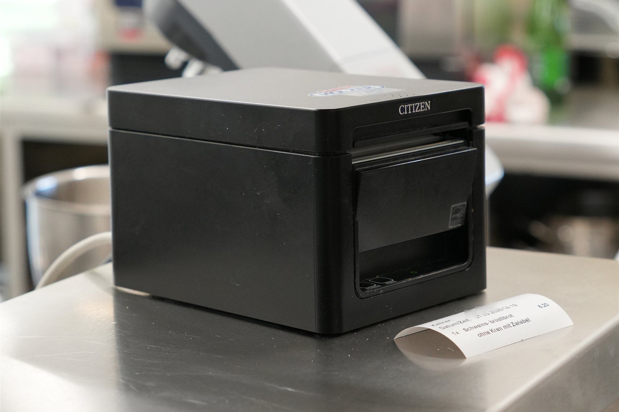 Bondrucker von Citizen auf der Edelstahlfläche einer Gastroküche, davor ein ausgedruckter Küchenbon mit einer Bestellung