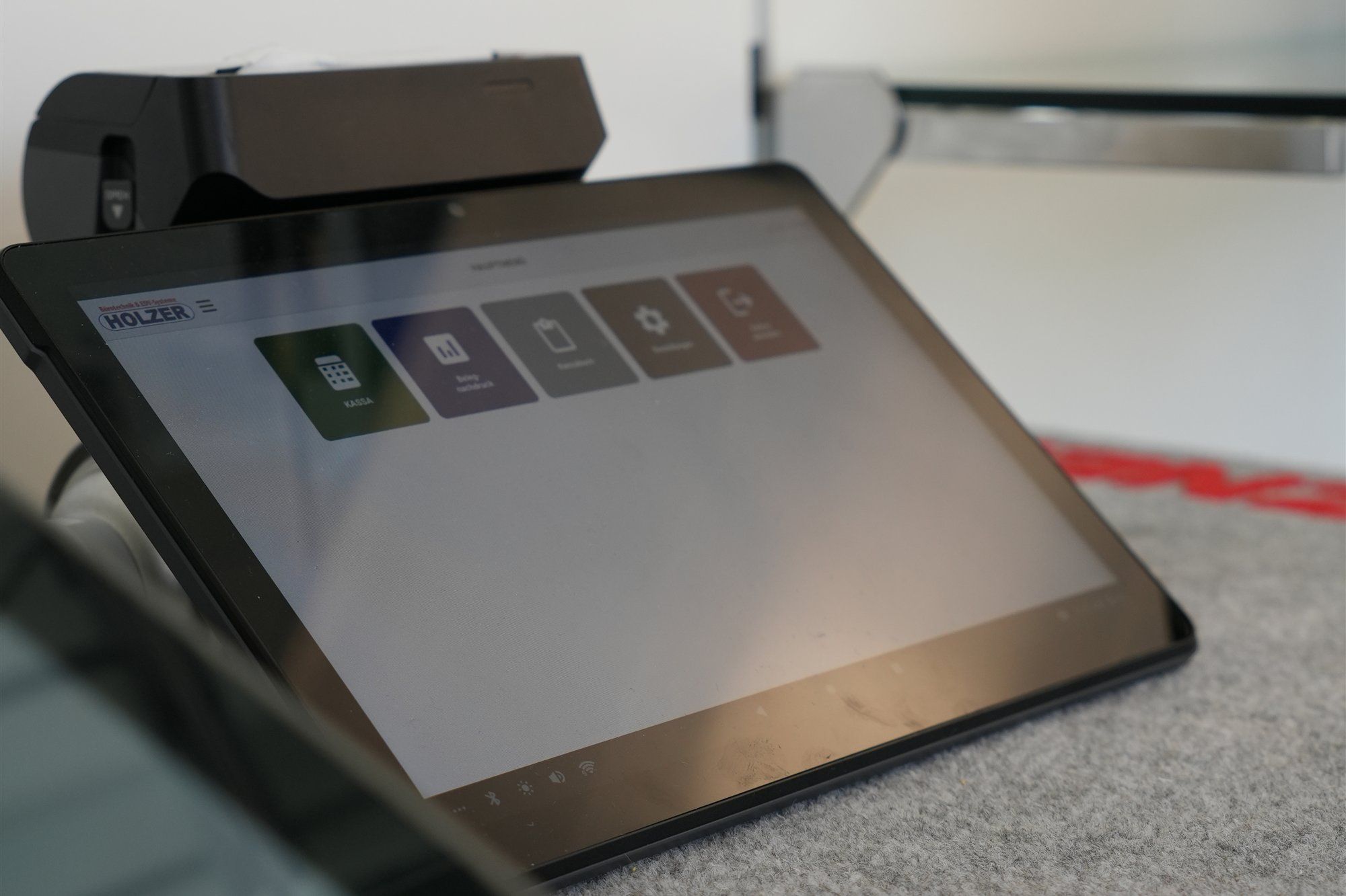 Touchscreen-Kassa mit dem Hauptmenü der Holzer-Kassensoftware, dahinter ein Bondrucker