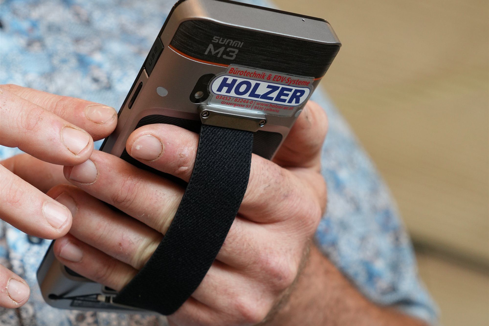 Hände halten ein Kassen-Handgerät von Sunmi mit Halteband und Holzer-Aufkleber