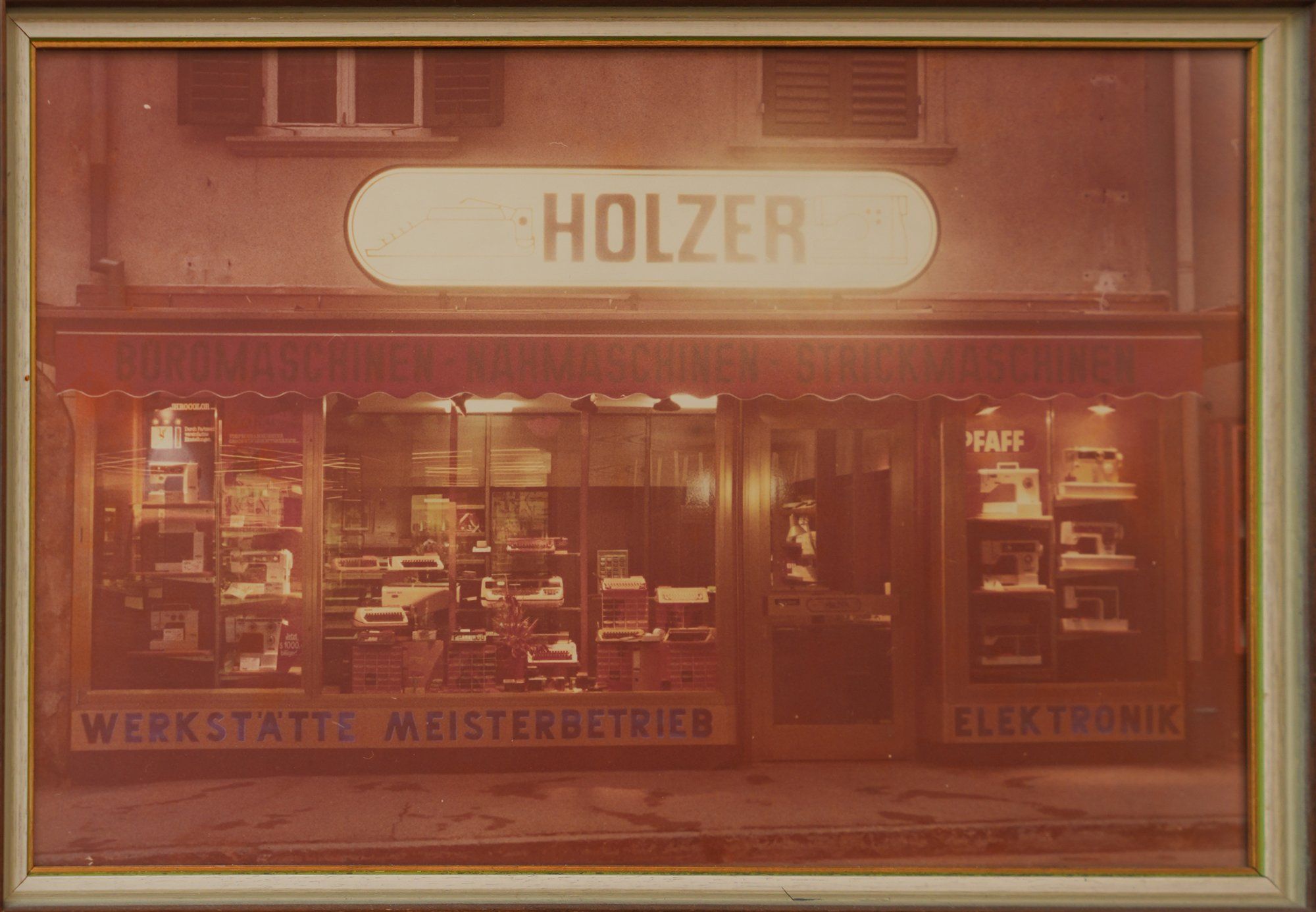 Gerahmte historische Aufnahme des Holzer-Geschäfts: Schaufenster mit Schreib- und Nähmaschinen, darüber die Aufschrift Büromaschinen, Nähmaschinen, Strickmaschinen