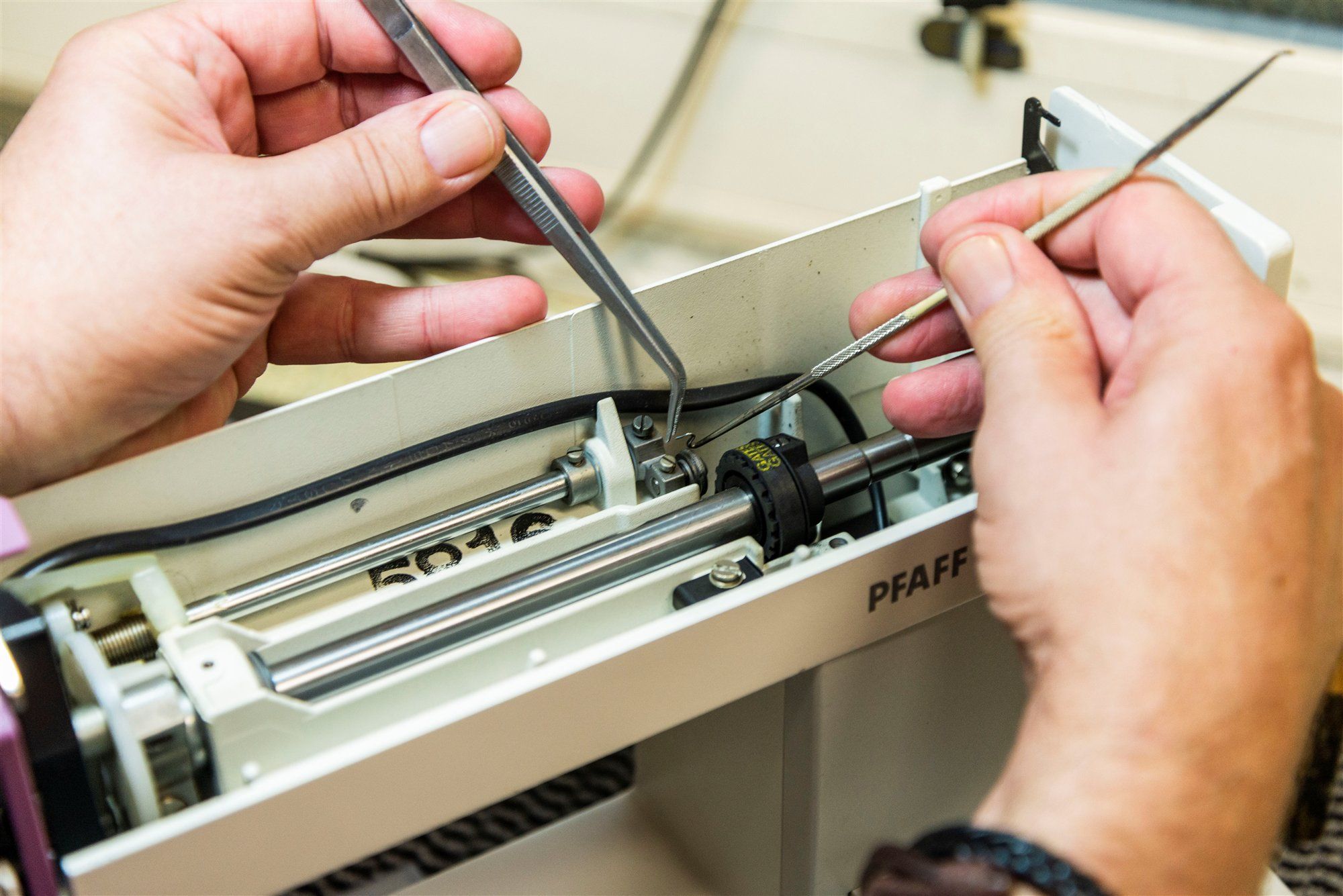 Hände eines Technikers mit Pinzette und Werkzeug an der geöffneten Mechanik einer Nähmaschine