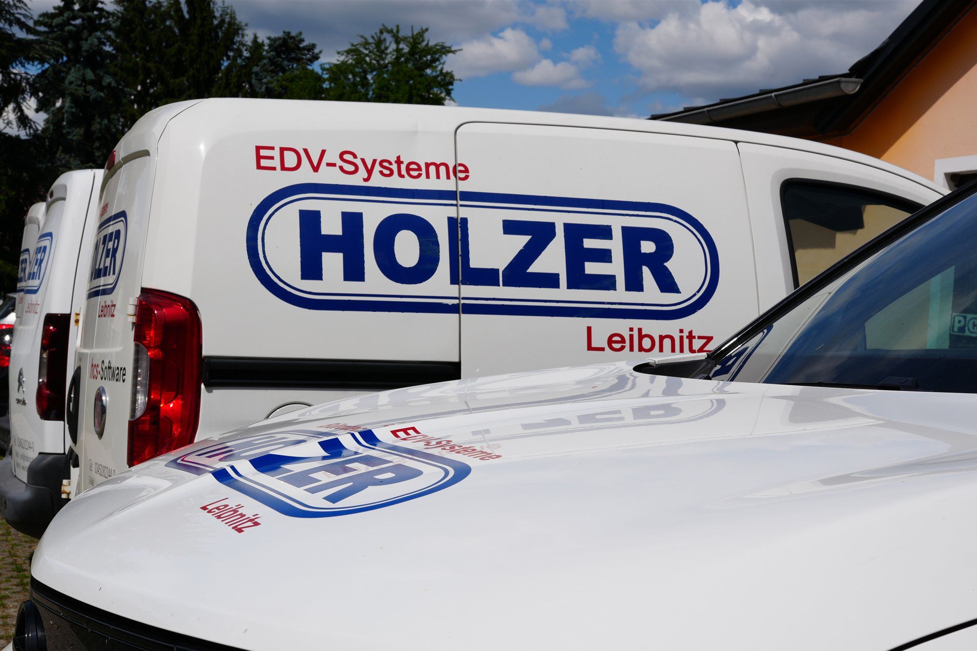 Weißer Lieferwagen mit großem Holzer-Logo und der Aufschrift EDV-Systeme Leibnitz, davor die Motorhaube eines Firmenwagens mit Logo